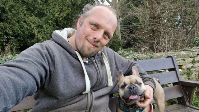 Dan and his dog sit on a bench outside a churchyard.