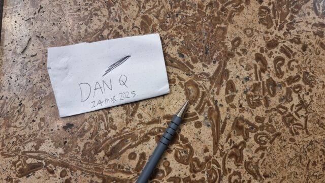 "Dan Q" and today's date written on a small piece of paper, alongside a pen, which points to a bivalve fossil in a limestone floor.