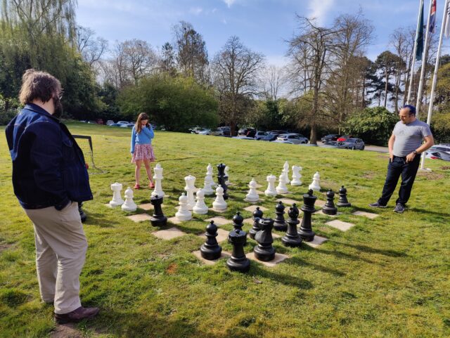 A girl studies a chessboard on a lawn, watched over by two men: Dan and JTA.
