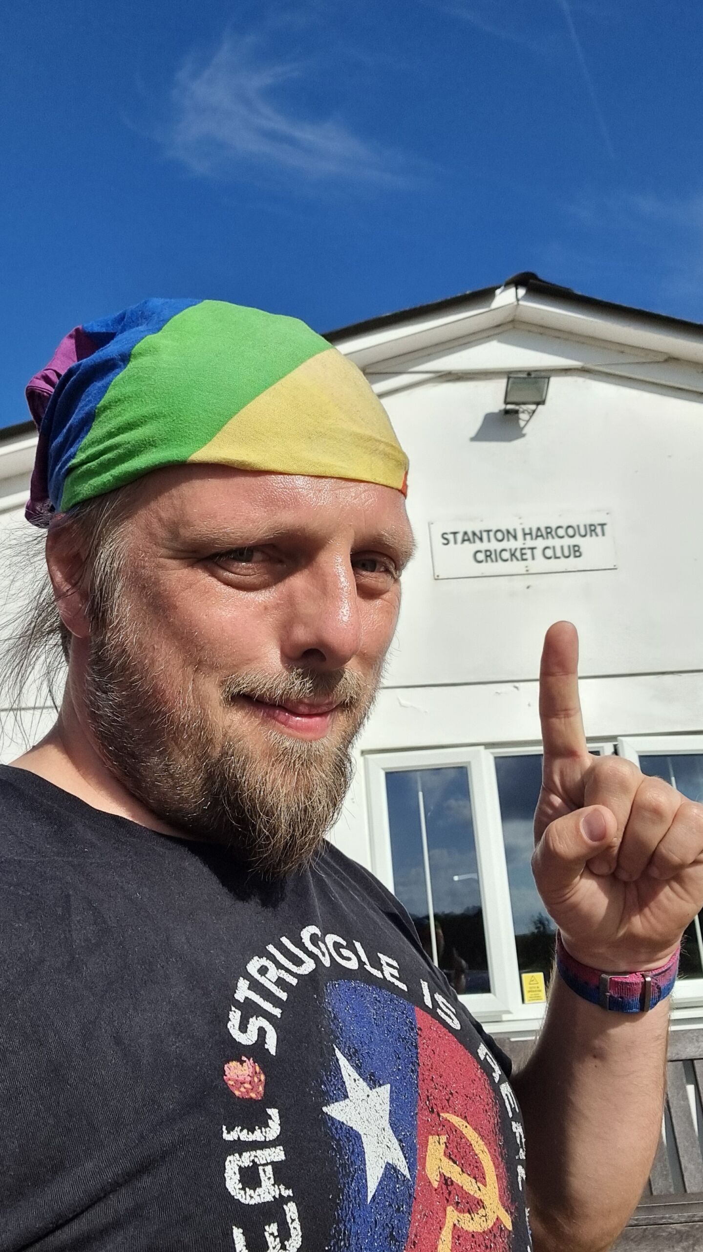 Larger image: Dan, a white man with a beard, wearing a rainbow bandana and a black t-shirt, points to a building in the background whose sign identifies it as Stanton Harcourt Cricket Club.