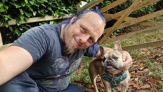 Dan, a white man with blue hair tied in a ponytail and a goatee beard, crouches alongside and pets a champagne-coloured French Bulldog by a gate on a path through a managed forest. There are bits of plant on his clothes, especially his shoulders.