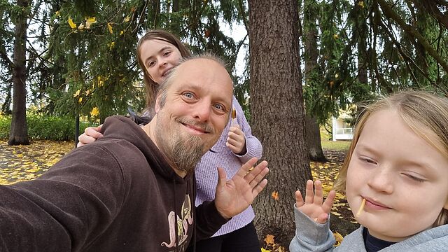 Dan and two children wave cheerfully from in front of a tree.