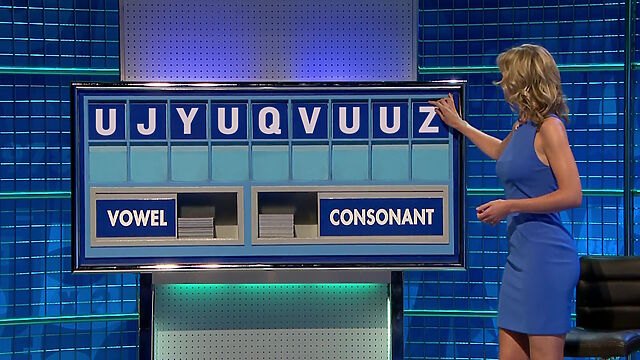 Rachel Riley, wearing a blue dress, places the last tile on a board to complete the tileset 'U J Y U Q V U U Z'.