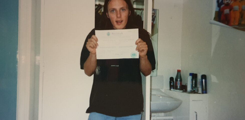 Standing in a study bedroom, Dan shows off his certificate of admission to the University of Wales, Aberystwyth.
