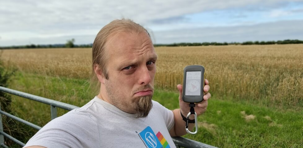 Dan, looking sad, holding a GPSr at a gate in front of a field of corn.