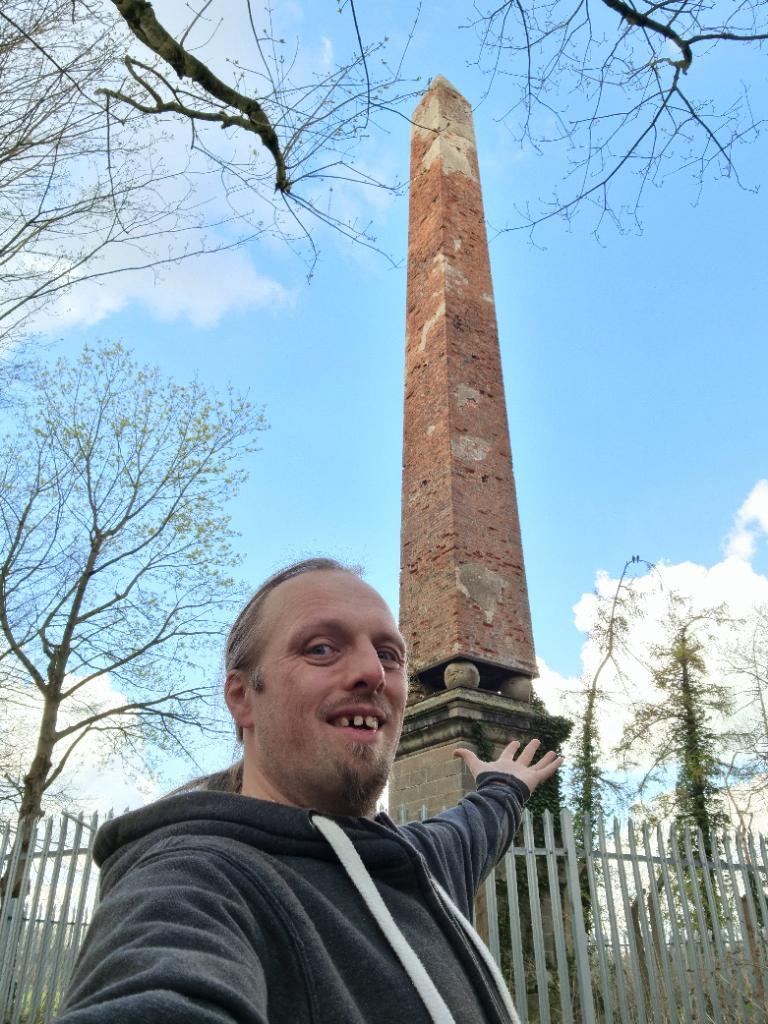 Dan stands under a brick obelisk under a mostly-clear blue sky. He is smiling and pointing at the landmark.