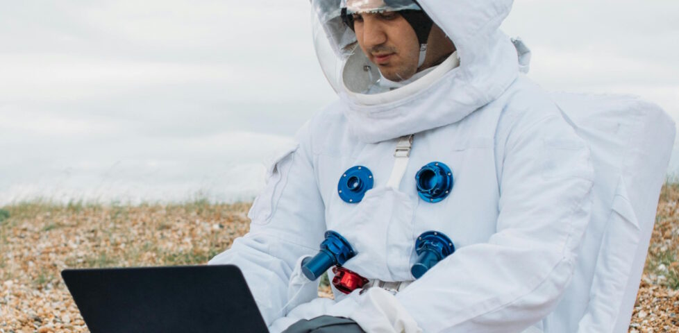 A white man wearing a spacesuit sits on a pebble beach using a laptop.