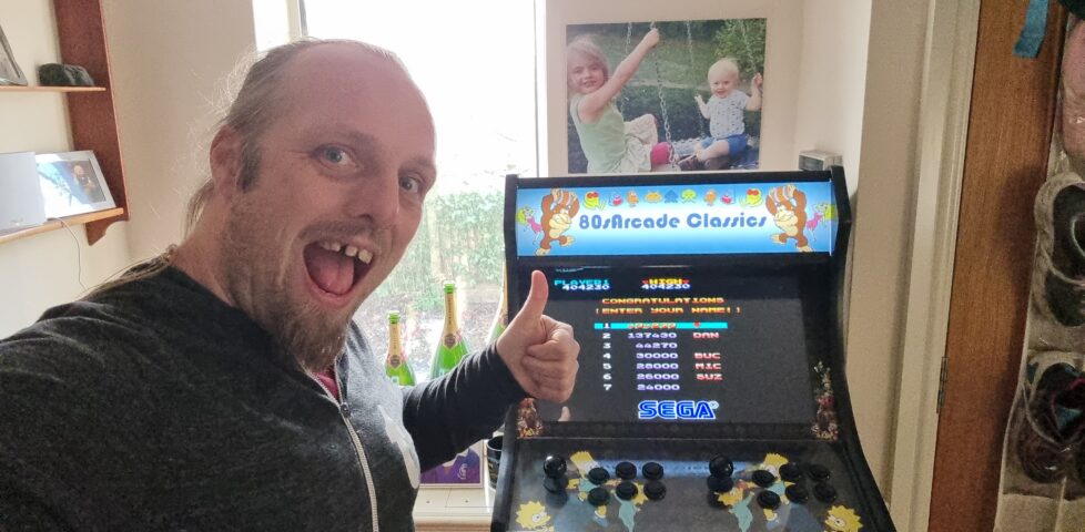 Dan with his thumbs-up in front of the high-score table (with the top-ranking spot about to be filled) of Wonder Boy, on a generic "80s Arcade Classics" arcade cabinet.