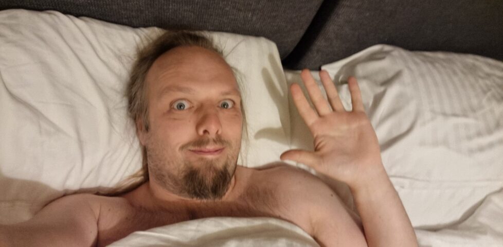 Dan waves, his head and shoulders peeping out from underneath a white duvet.