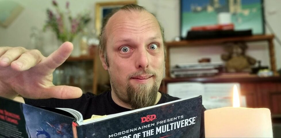 Dan gestures with a wave as he peers into the camera over the top of a copy of Monsters of the Multiverse, by candlelight.