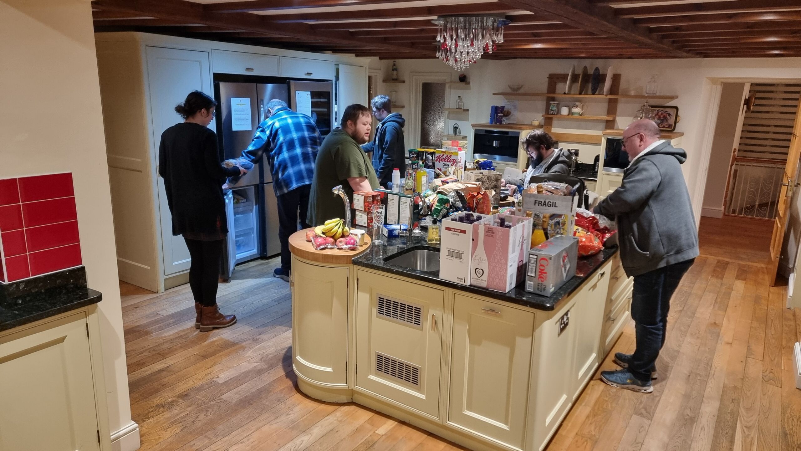 Full image of A large group of people stand around a pile of shopping in the centre of a nicely decorated kitchen.