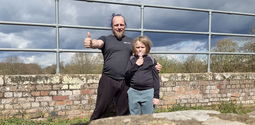 Dan and a small child throw a thumbs-up to the camera from atop a brick bridge.