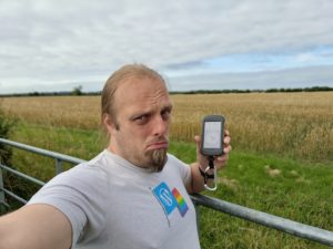 Dan, looking sad, holding a GPSr at a gate in front of a field of corn.