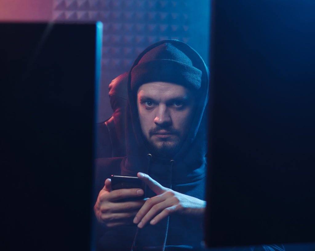 "Hacker type" man in hoodie between two computer monitors, looking at his phone.