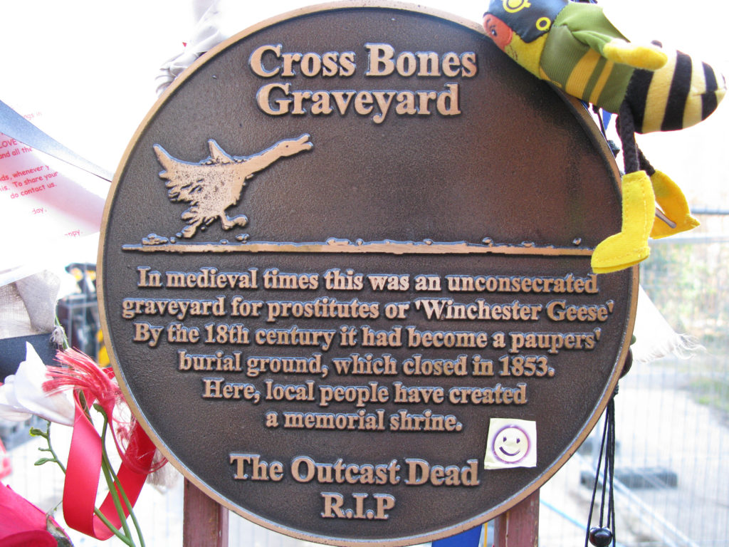 Plaque with a picture of a goose running and text: "Cross Bones Graveyard. In medieval times this was an unconsecreated graveyard for prostitutes of 'Winchester Geese'. By the 18th century it had become a paupers burial ground, which closed in 1853. Here, local people have created a memorial shrine. The Outcast Dead R.I.P." A smiley face sticker has been attached to the plaque and ribbons and silk flowers are tied nearby.
