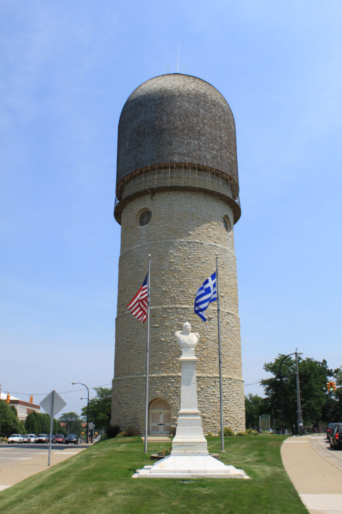 The Ypsilanti Water Tower, at the intersection of Washtenaw Avenue and Cross Street, Ypsilanti, Michigan. The tower is listed in the National Register of Historic Places, and is a National Historic Civil Engineering Landmark. An American flag and a Greek flag are flying, and a bust of the Greek general, Demetrios Ypsilantis (also commonly spelled "Demetrius Ypsilanti"), for whom the city is named, is in the foreground. Photo by Dwight Burdette, used under a Creative Commons license.