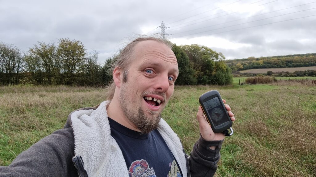 Dan makes a silly grin while holding a GPS receiver in a damp grassy autumn meadow, on a grey and drizzly day.