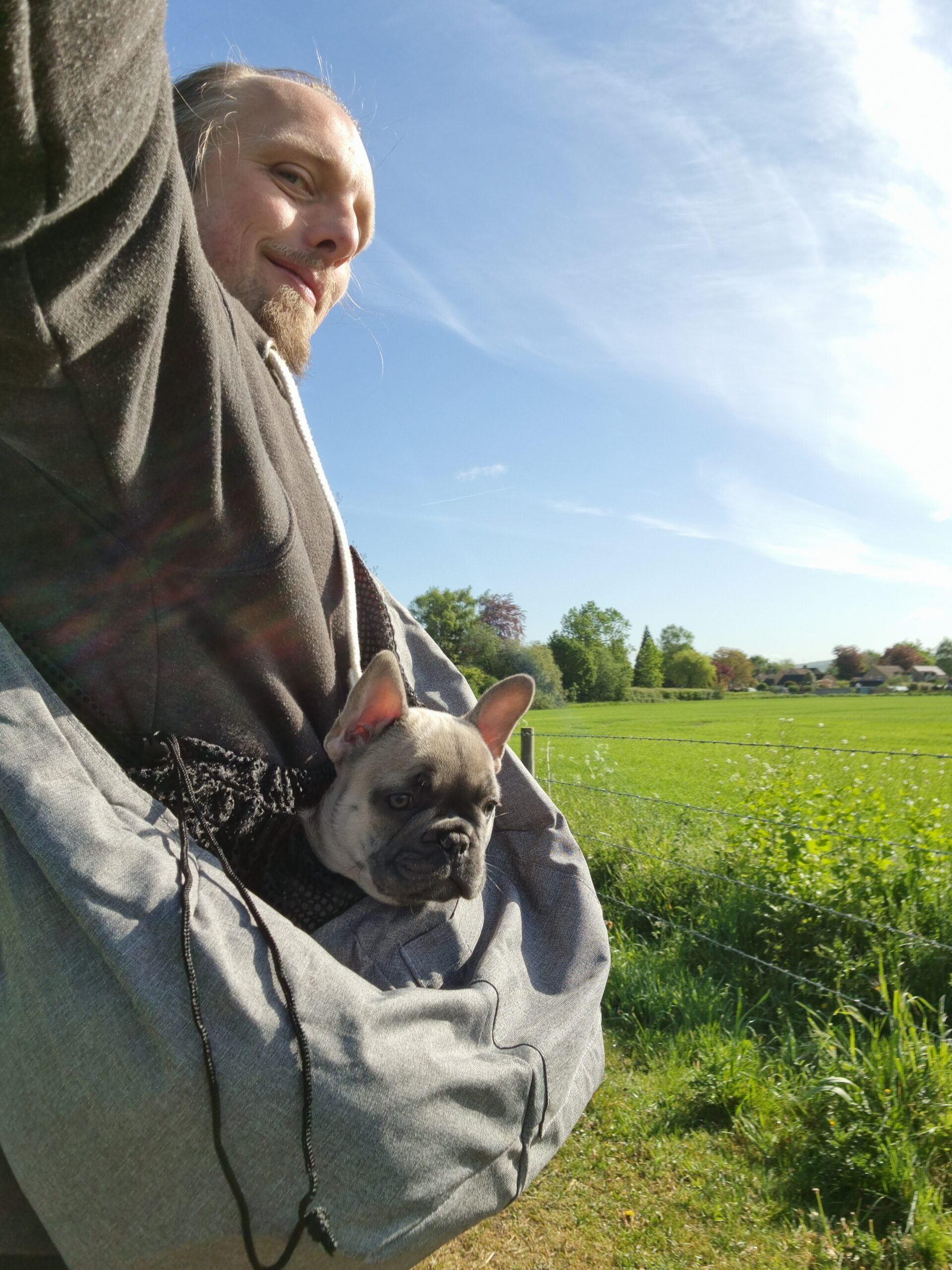 Dan carrying French Bulldog 'Demmy' in a dog carrier bag, with a sunny meadow behind them.
