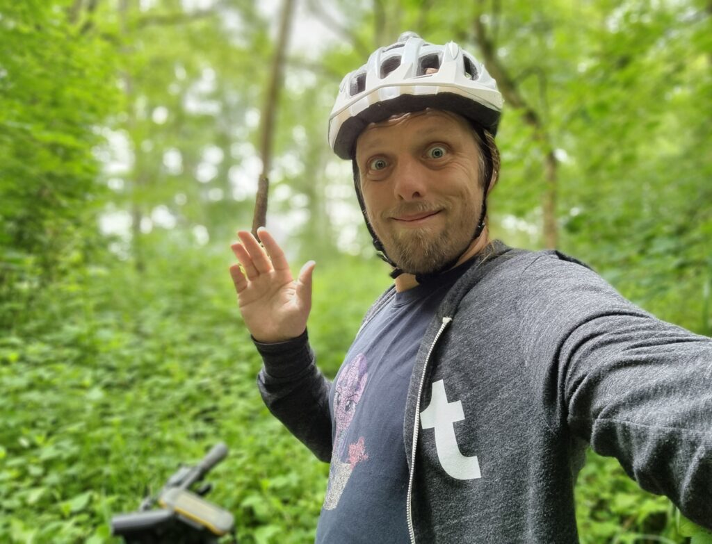 Dan, wearing a Tumblr hoodie over a My Little Pony T-shirt and with a white cycle helmet on his head, stands in front of his bike (with a GPS receiver on the handlebars) in a deep green forest.