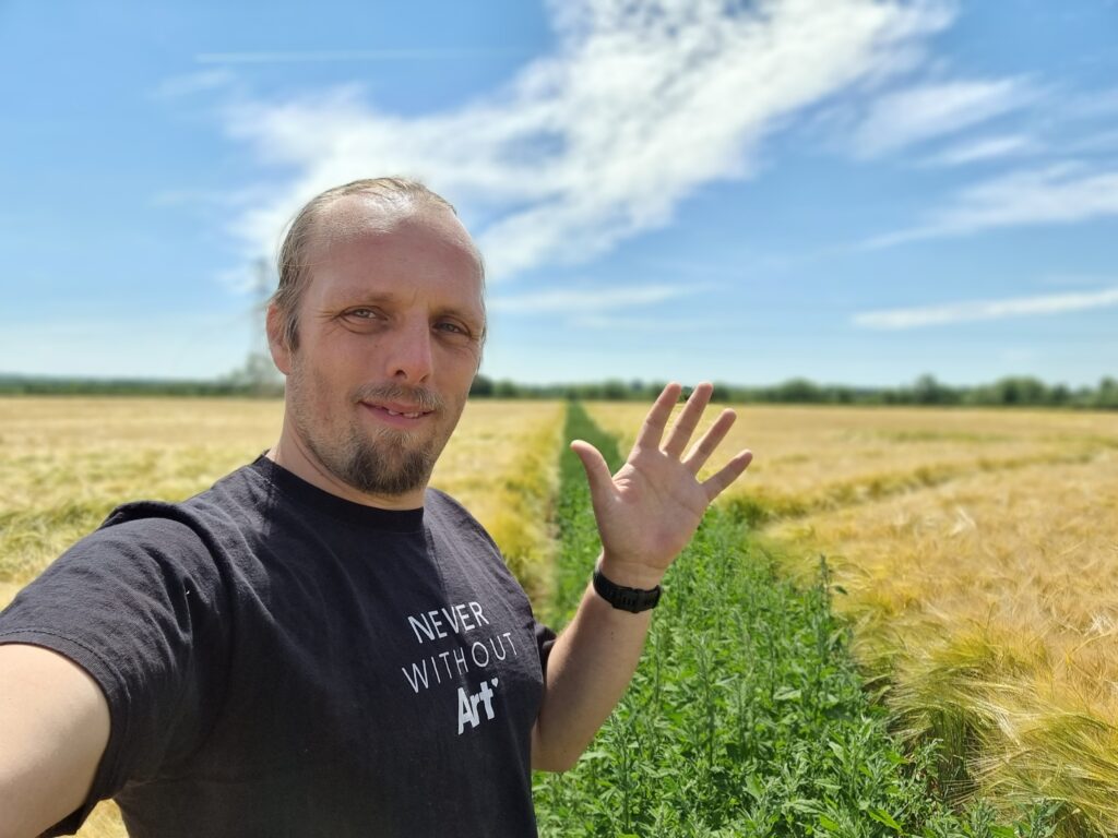 Dan, in a black t-shirt with "never without art" written on it, waves to the camera. He's standing in a cornfield, but the "path" that cuts diagonally through it is filled with plants to a similar height as the corn, making it hard to pass.
