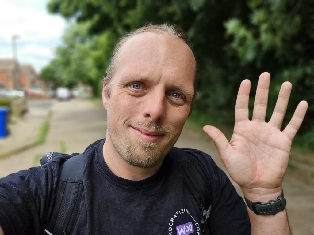 Dan, in a black t-shirt, waves to the camera from a suburban street.