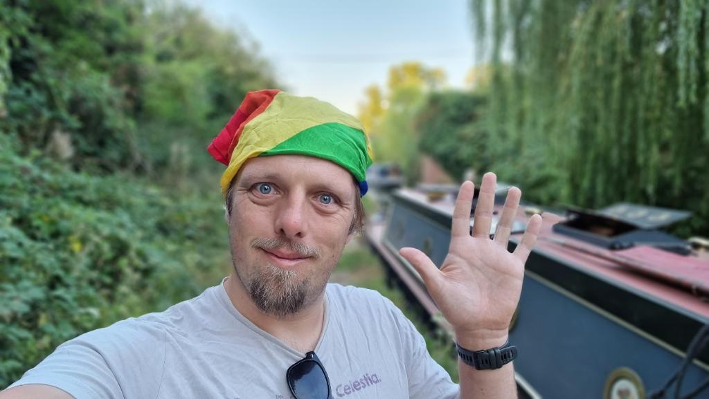 Dan, wearing a rainbow-striped bandan, waves to the camera from a rural canal towpath.