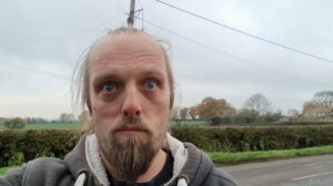 Standing in front of an advancing cloud front in a rural village, Dan - dressed in a grey and white Three Rings hoodie - stares sadly into the camera.