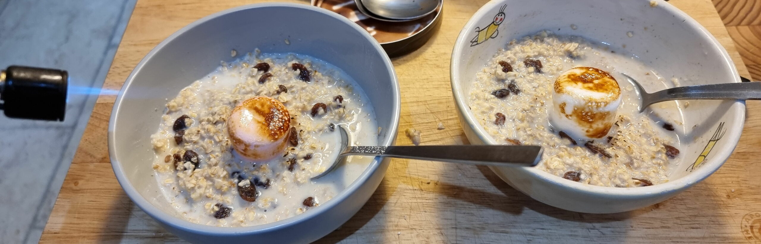Two bowls of porridge with sultanas, each topped with a jumbo marshmallow (one pink, one white) being toasted by a blowtorch (mostly off-camera).