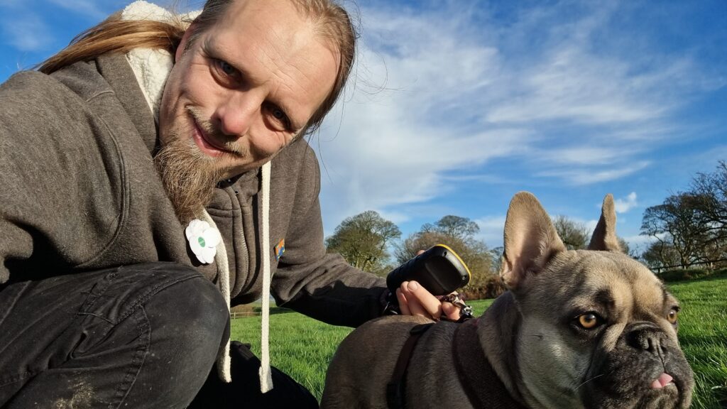 Dan - a man with a beard, wearing a grey fleece with a white poppy attached - crouches alongside his French Bulldog in a green field under a blue sky with a few wispy clouds.
