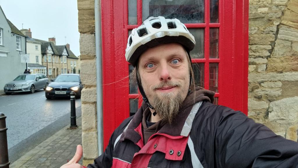 Dan, wearing a red-and-black jacket and a white cycle helmet, stands in front of a red telephone box on a village street.
