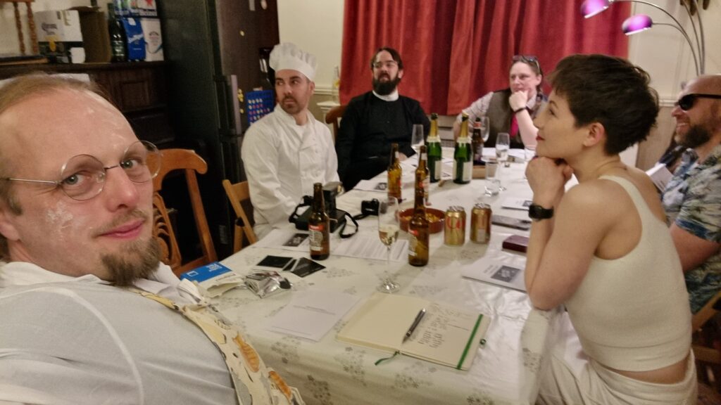 A group of people sit around a dining table; one man, wearing spectacles on his floury face, is taking the photo in "selfie" mode. The other visible characters appear to be a chef, a vicar, a librarian, a thrillseeker, and an aspiring starlet. With the exception of the photographer, they're all looking off to the side, listening to an unseen character speak.