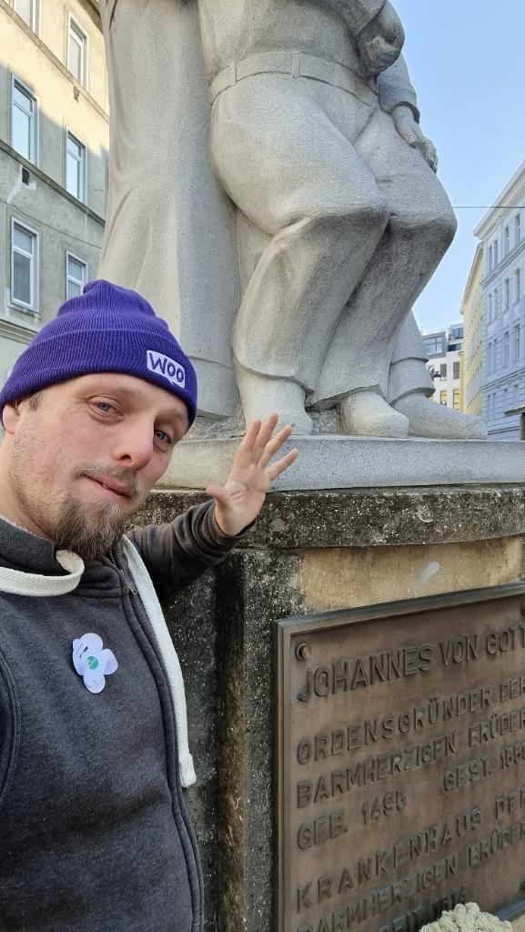 Dan, wearing a purple "Woo" wooly hat, stands in front of the base of a statue of Johannes von Gott.