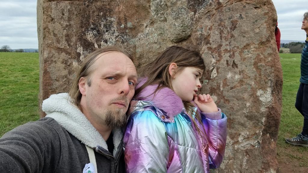 Dan, in a grey hoodie, stands with a nine-year old girl in an iridescent coat. The pair have their ears pressed against a tall standing stone as if listening to it.