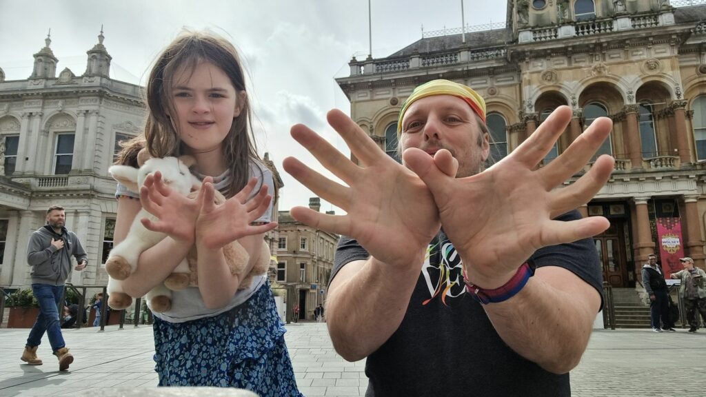 Dan, wearing a black t-shirt and a rainbow-striped bandana, stands alongside a 10-year old girl carrying stuffed toys, in a market square. Both are making "spider" figures with their fingers, towards the camera.