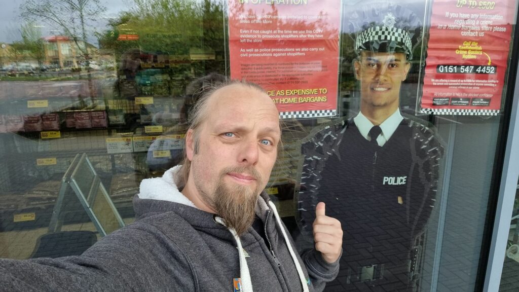 Dan stands outside a floor-to-ceiling shop window within which is a cardboard cut-out of a smiling police officer.