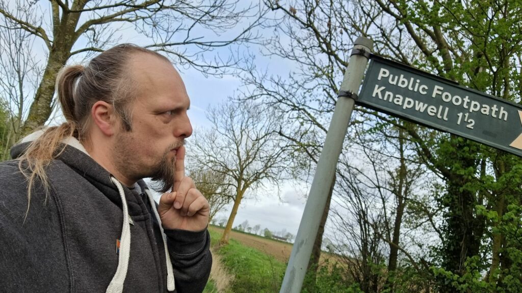 Dan, his finger to his lips as-if in thought, looks at a finger post sign that indicates a public footpath to Knapwell (1½ miles).