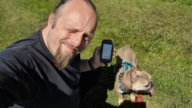 Dan squints into the sunlight in a grassy field, alongside a dog.