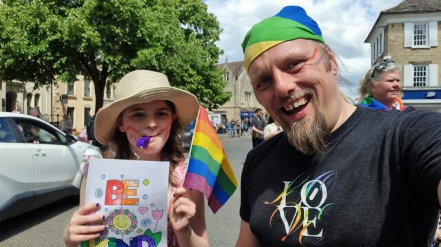 A tweeny girl and a 40-something man with rainbows painted on their faces wave flags in a Pride parade. The child has coloured-in a poster saying "be kind".