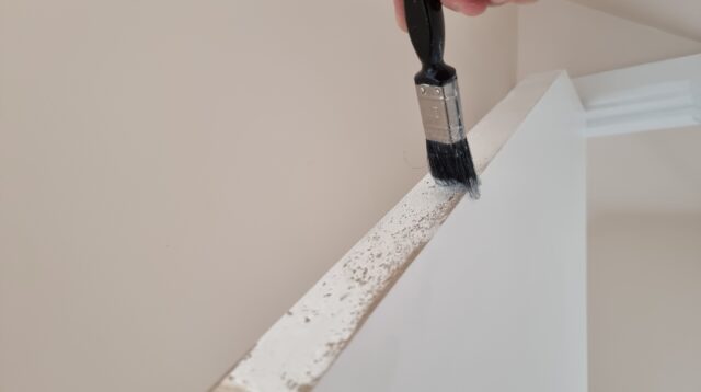 A paintbrush applies white paint to the top of a door.