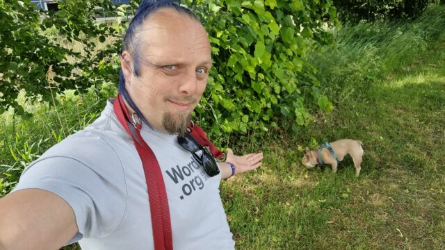 Standing in a green park space, Dan gestures to his dog, a Frenchie, who's sniffing a nearby tree.